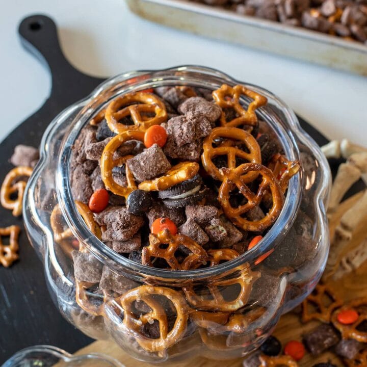 Halloween Muddy Buddies with chocolate-coated Rice Chex, mini pretzels, Oreos, and orange and black M&M's in a glass pumpkin jar.